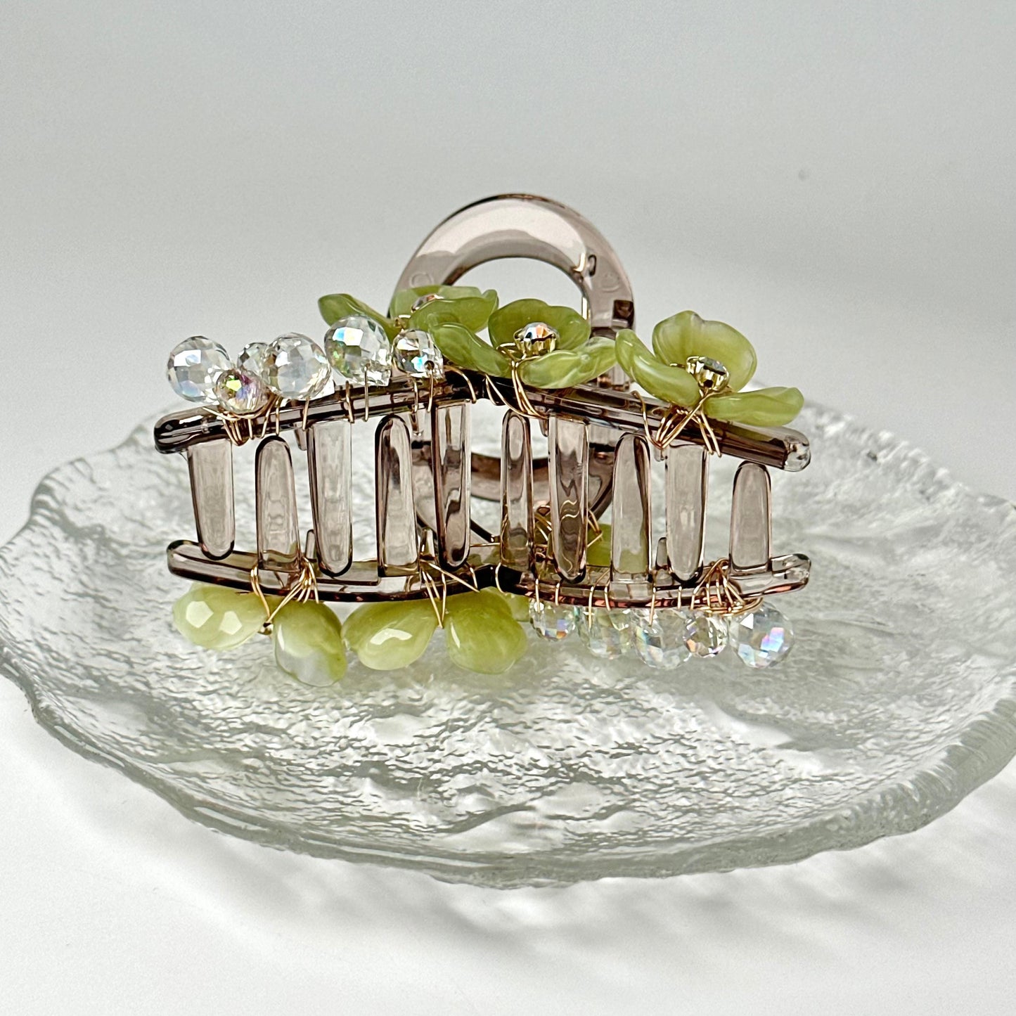 A clear glass plate with a decorative hair clip featuring green and clear gemstone accents.
