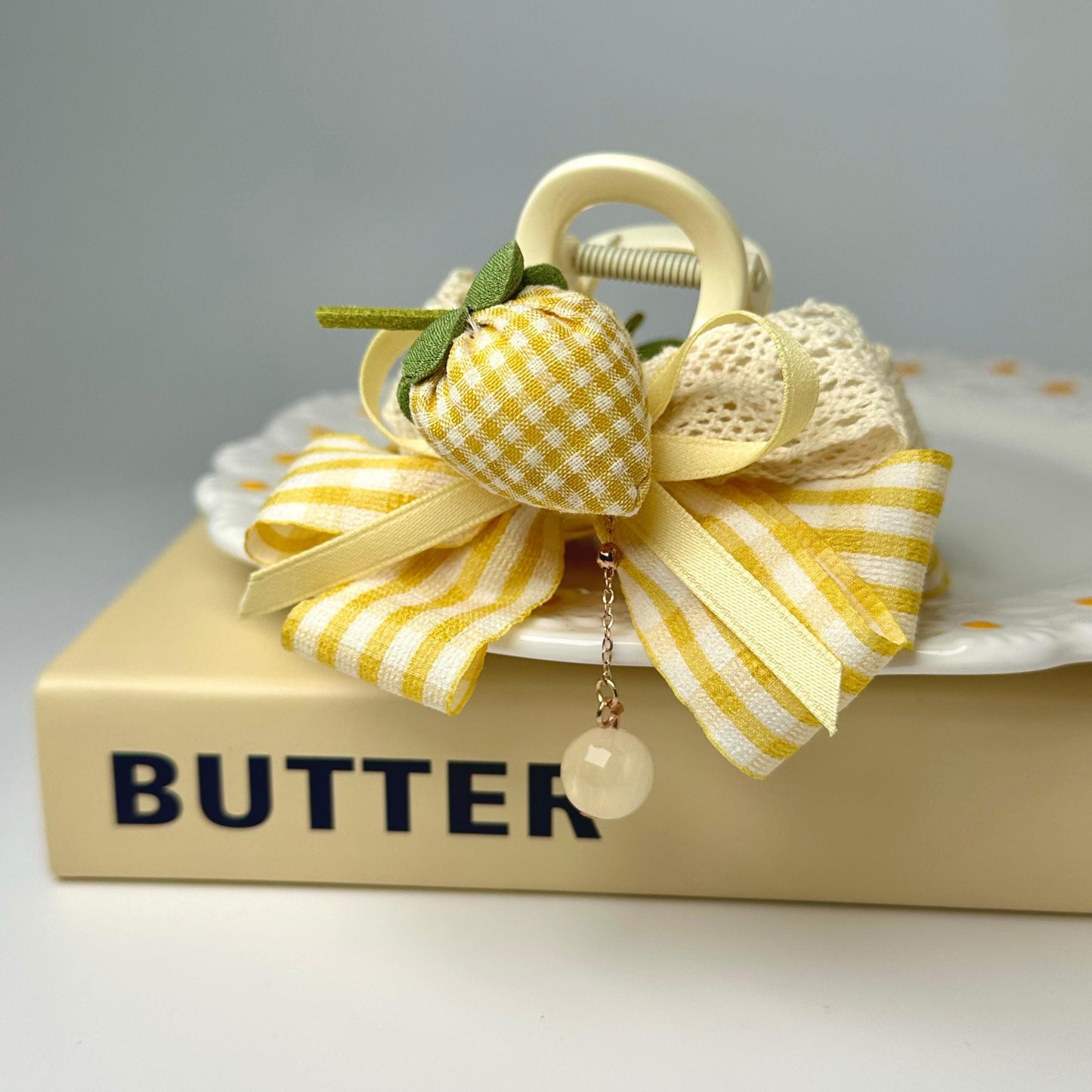 A decorative strawberry-shaped hair clip with a yellow and white striped ribbon sits on top of a book titled "BUTTER".