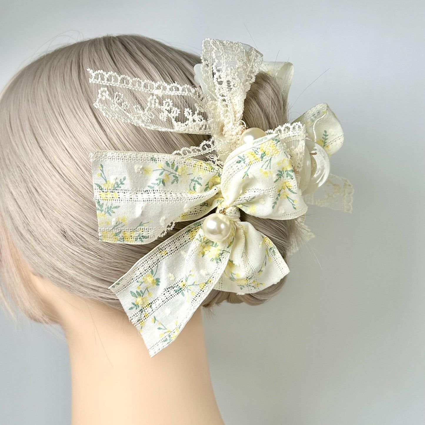 A mannequin head with a blonde hair styled in a bun, wearing a decorative hair accessory featuring a floral pattern and a pearl accent.