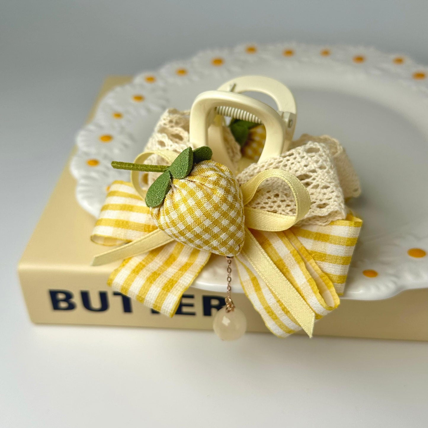 A decorative bow made of fabric and ribbons, featuring a yellow and white checkered pattern, is placed on top of a white plate with a scalloped edge.