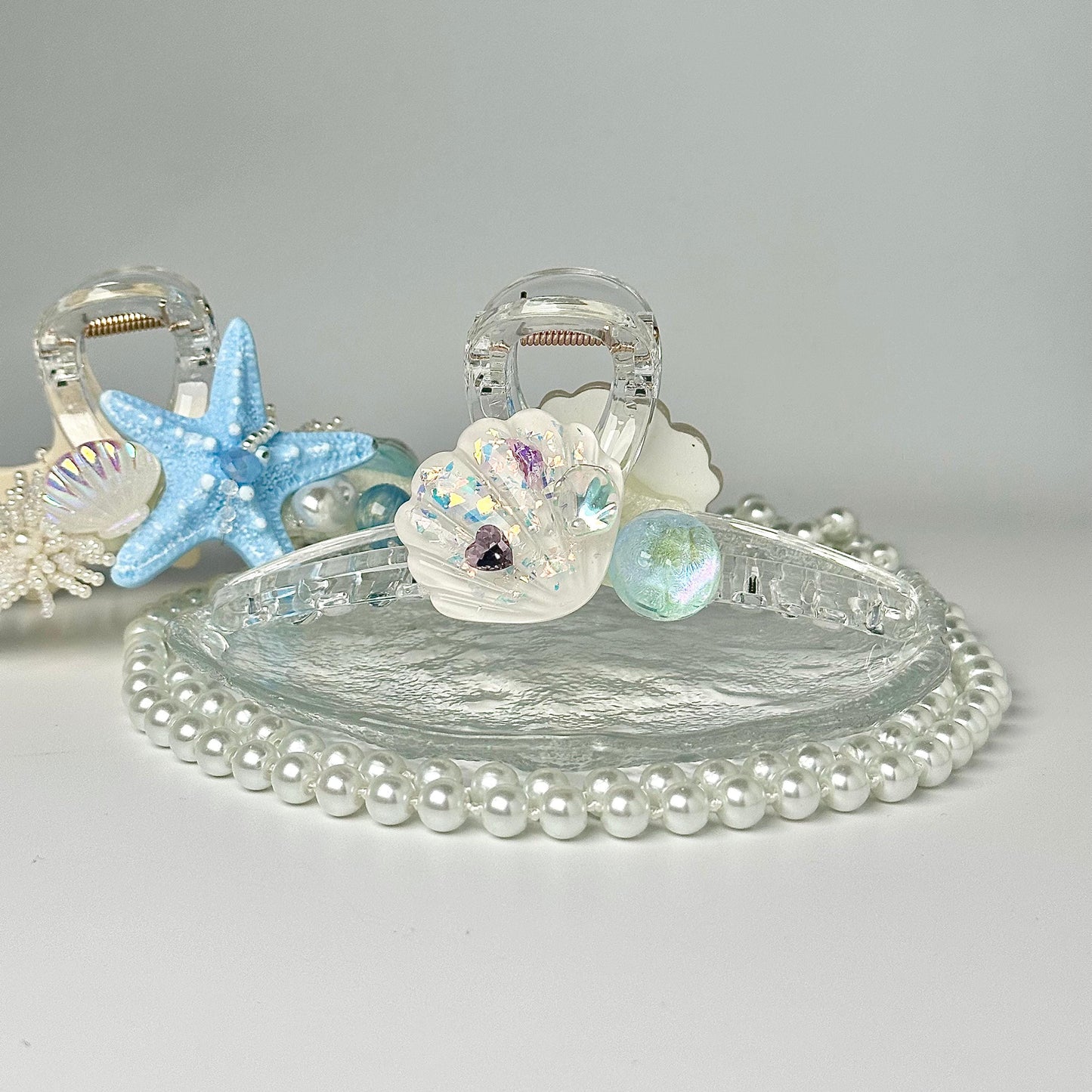 A clear glass hair clip adorned with seashells, pearls, and a starfish, set against a white background.