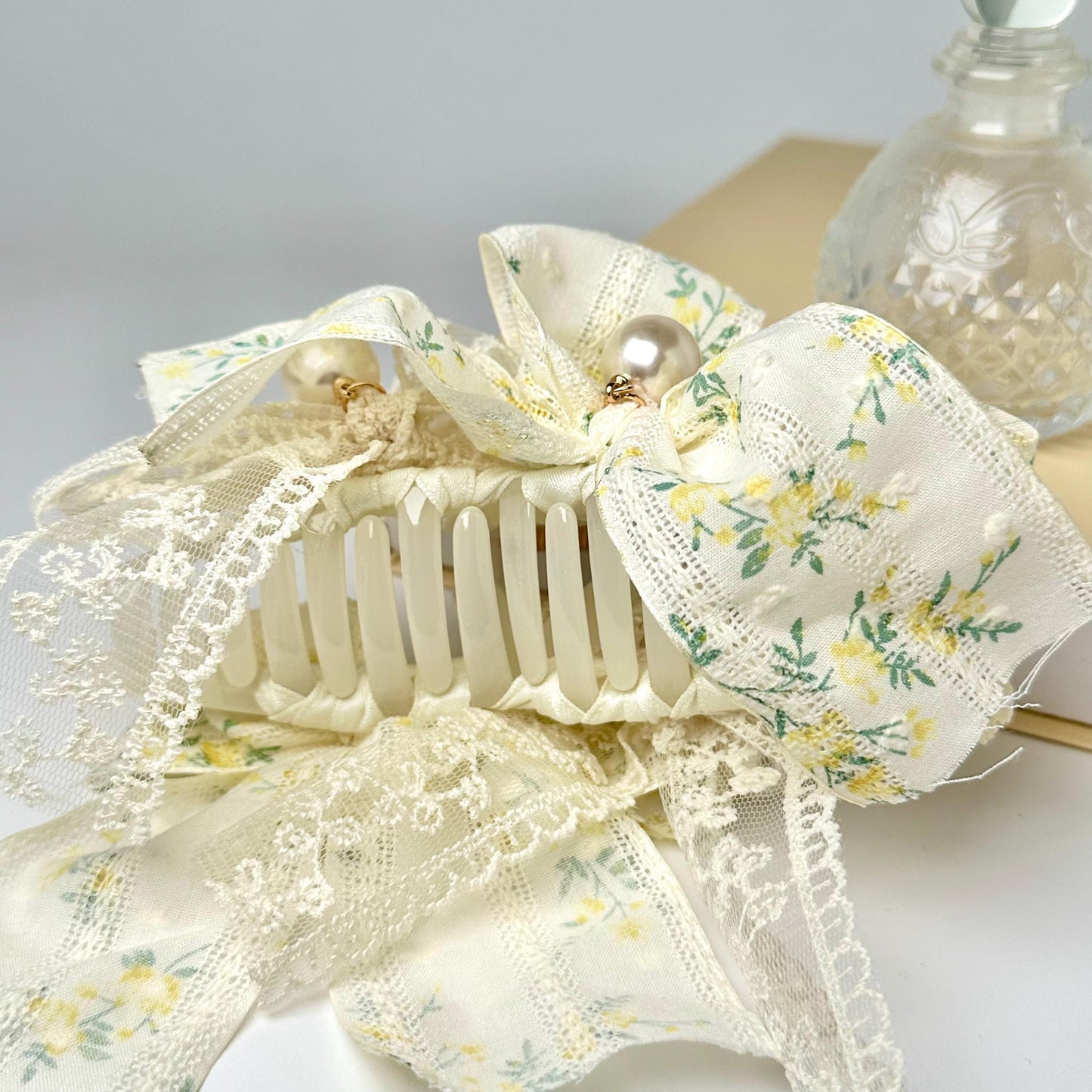 A delicate white lace ribbon with a floral pattern, tied into a bow, is displayed alongside a clear glass perfume bottle.