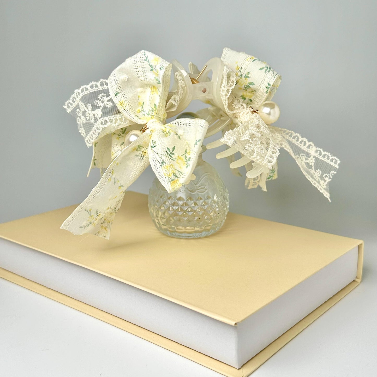 A clear glass vase filled with white flowers and lace sits on top of a yellow book, creating a charming and elegant display.