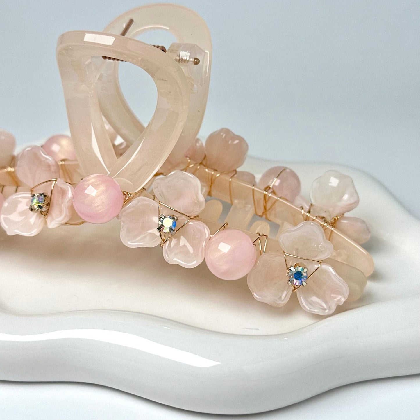 A close-up image of a delicate hair accessory, possibly a hair clip or headband, adorned with small, pink flowers and rhinestones or crystals.