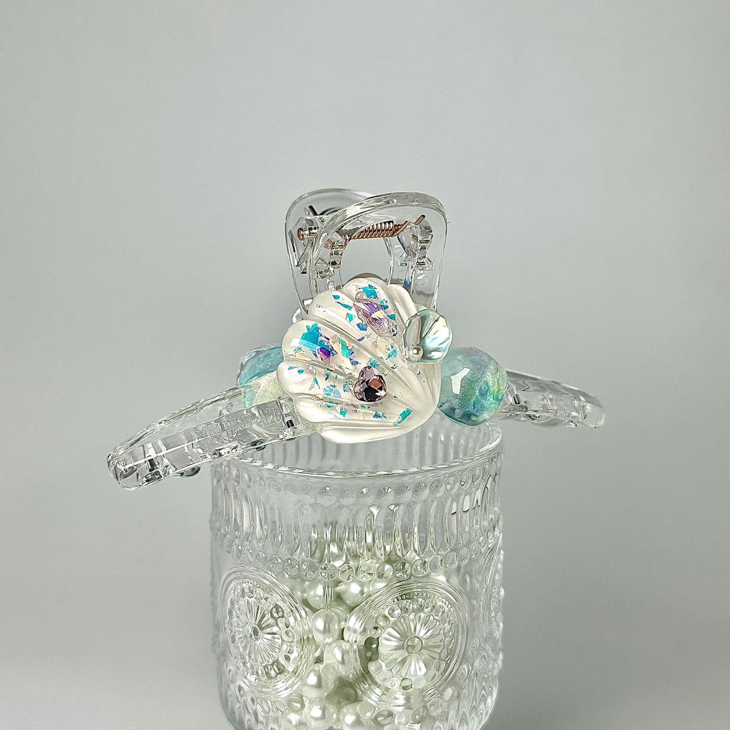 A clear glass container filled with small, round, multicolored pearls, with a clear plastic object featuring a shell-shaped design and colorful accents placed on top of the container.