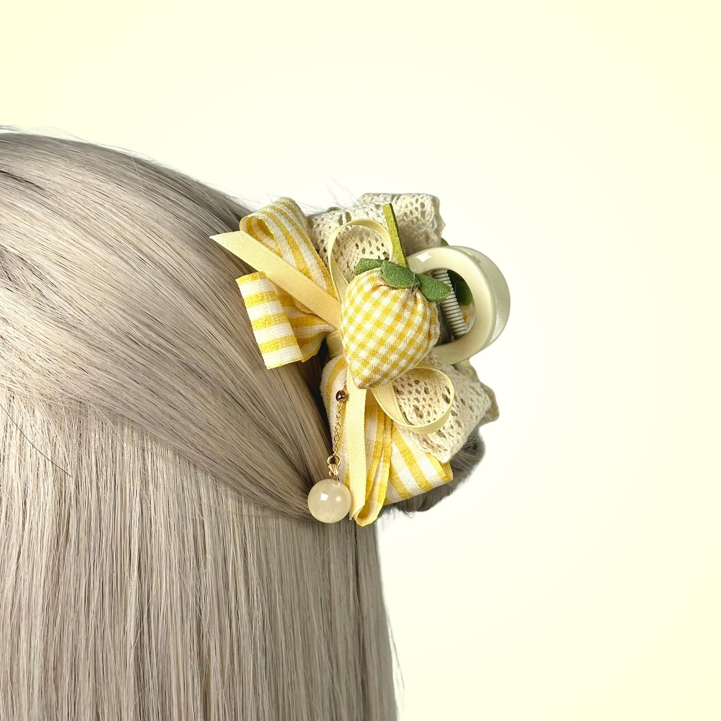 A close-up of a person's blonde hair with a decorative hair accessory featuring a yellow and white striped ribbon, a green leaf, and a pearl.