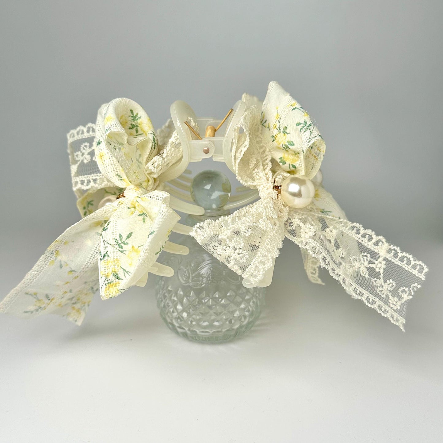 A glass vase with a floral pattern and lace trim, adorned with delicate bows and pearls, sits on a white surface.