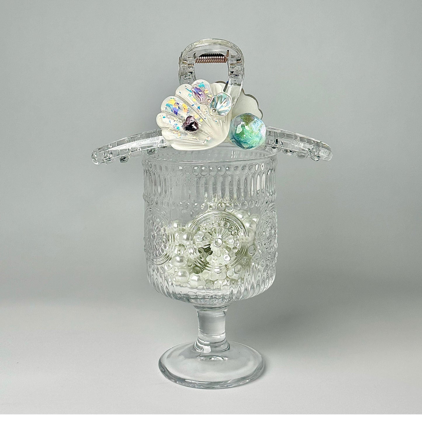 A clear glass vase filled with white pearls and adorned with a decorative shell-shaped clasp and colorful gemstones.