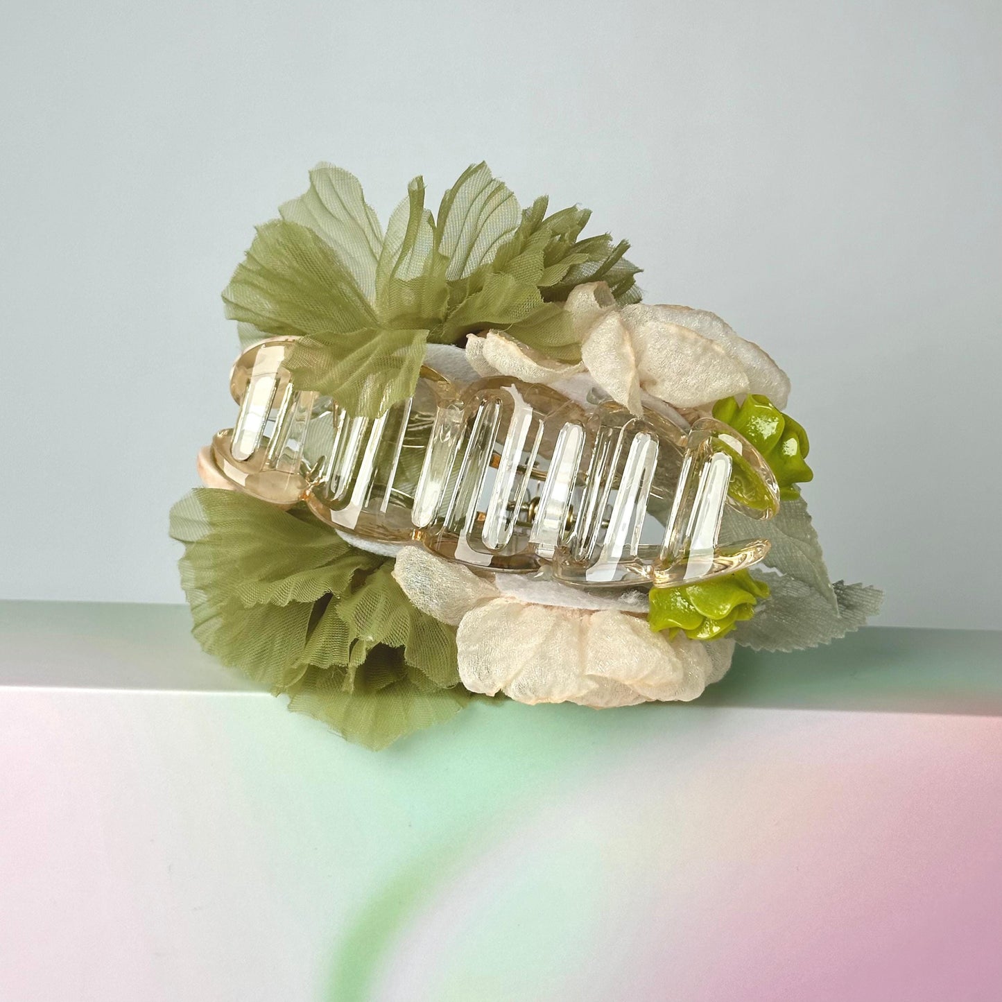A clear hair clip adorned with green and white flowers rests on a pink and green surface.