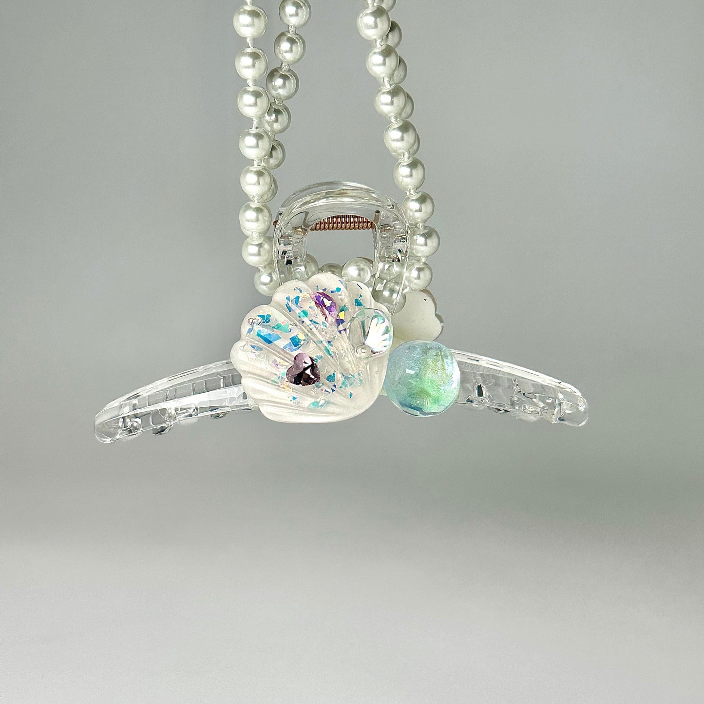 A necklace with clear beads and a shell-shaped pendant, adorned with colorful gemstones, is suspended against a light gray background.