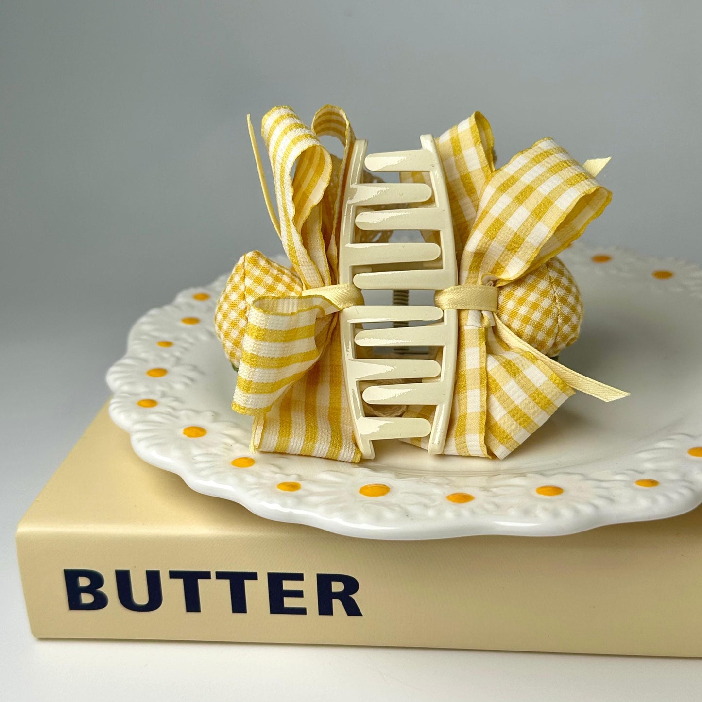 A white plate with a yellow and white checkered pattern and a white ribbon tied into a bow sits on top of a yellow book titled "BUTTER".