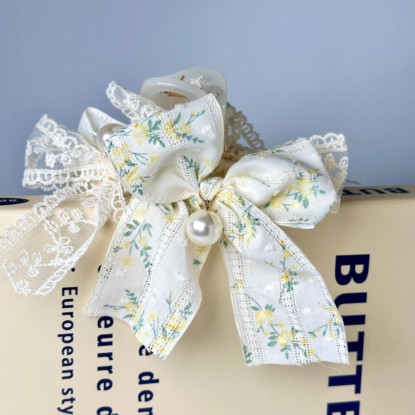 A close-up image of a white ribbon with a floral pattern tied around a white lace bow, placed on top of a book with the title "BUTTER" visible.