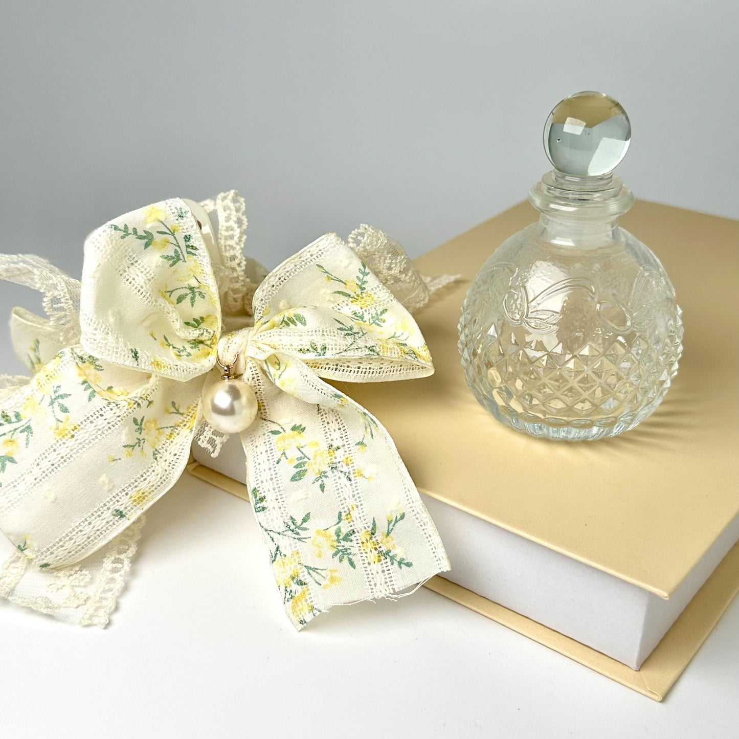 a clear glass perfume bottle with a white lace bow and a white book with a gold ribbon on top, all placed on a white surface.