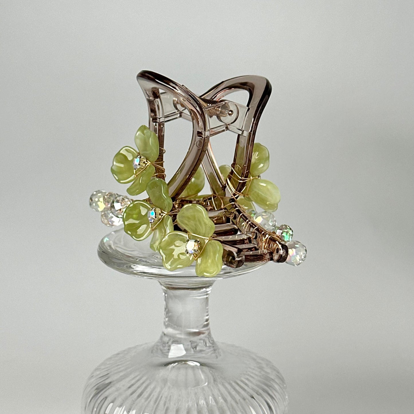 A clear glass vase holding a hair clip with green and silver flowers and crystals.