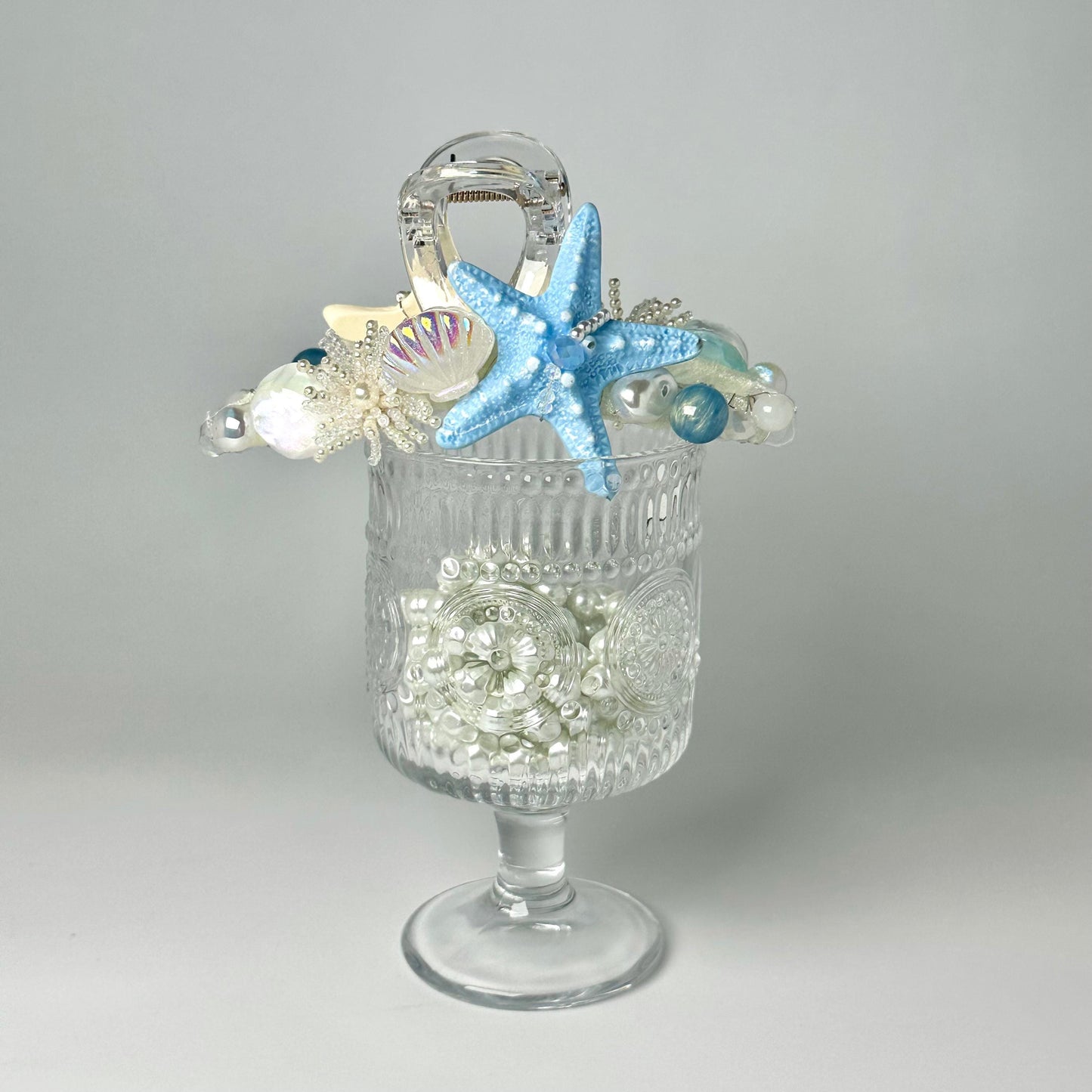 A clear glass vase filled with small white pearls and a blue starfish is placed on a white surface.