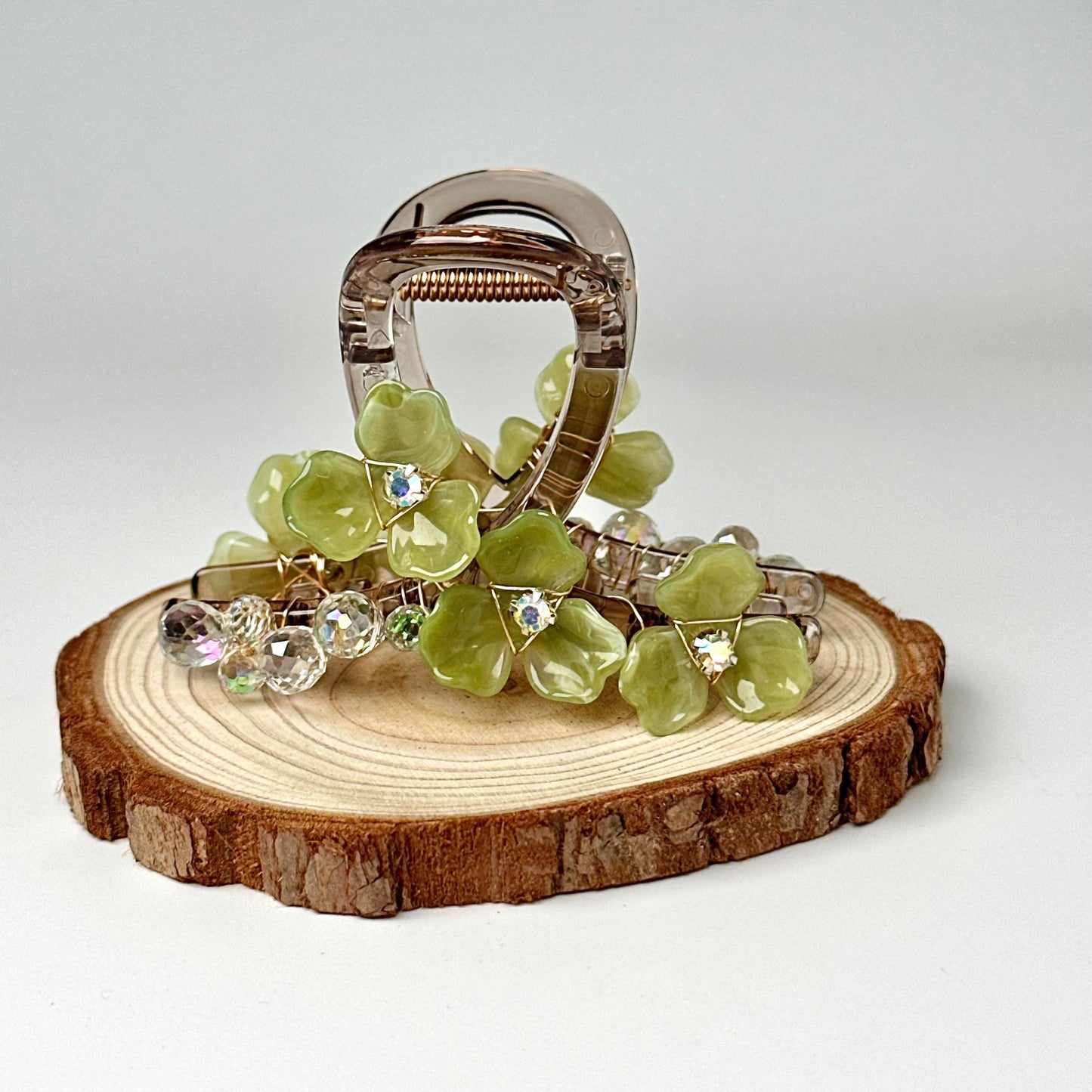 A silver hair clip adorned with green glass flowers and rhinestones is displayed on a wooden slice.