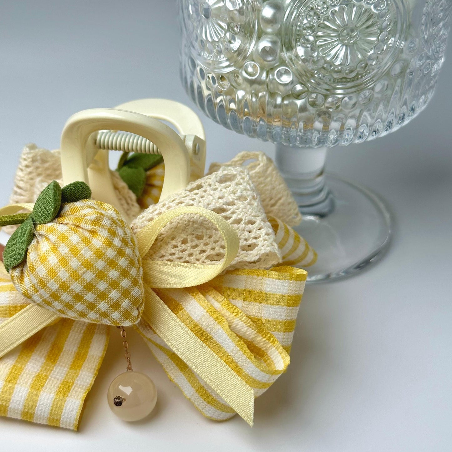 a close-up view of a decorative item, which appears to be a ribbon or bow, adorned with a small charm or bead. The ribbon is tied in a bow shape, and there is a glass vase or container with a floral pattern on the side, adding an elegant touch to the overall presentation.