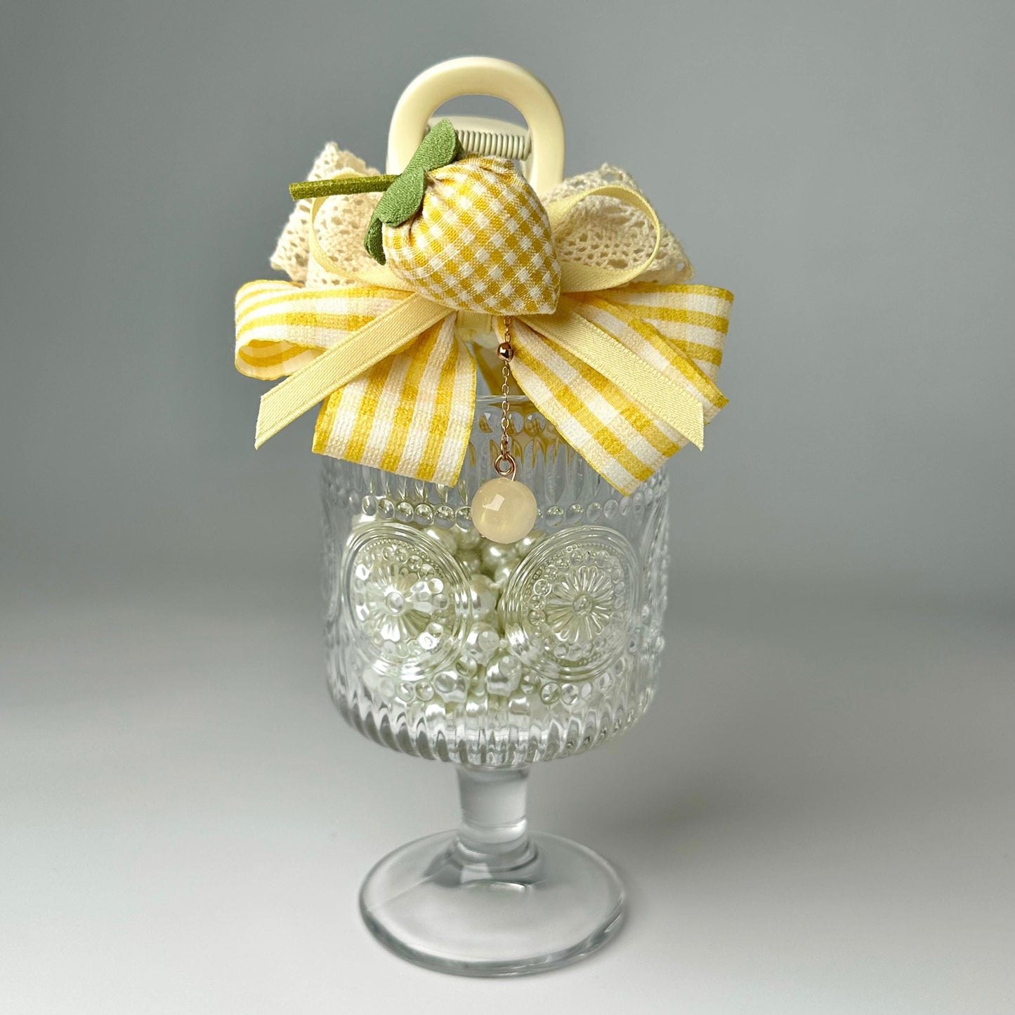 A glass vase with a yellow and white checkered bow, a small charm, and a white flower, placed on a white surface.
