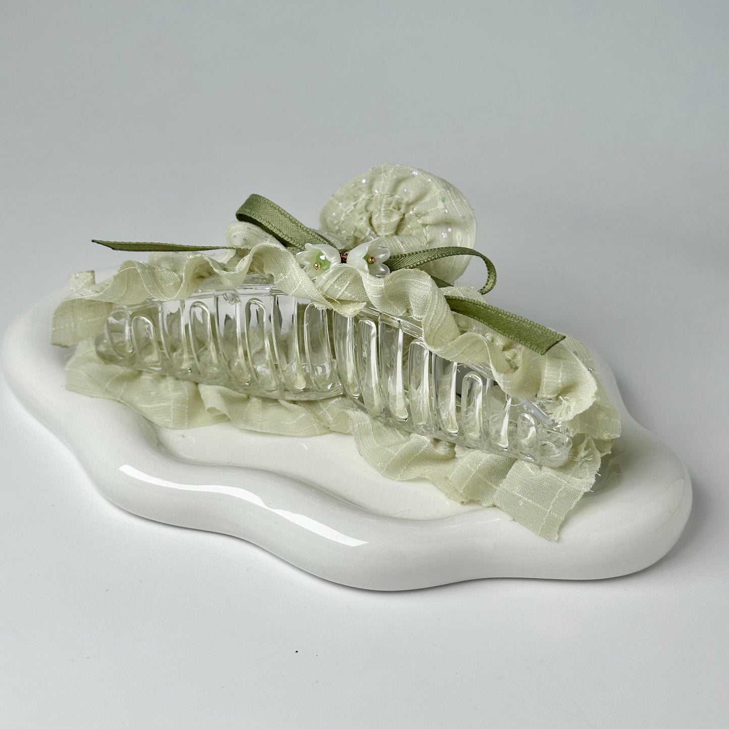 A clear glass hair accessory with a green ribbon and a white flower decoration is displayed on a white surface.