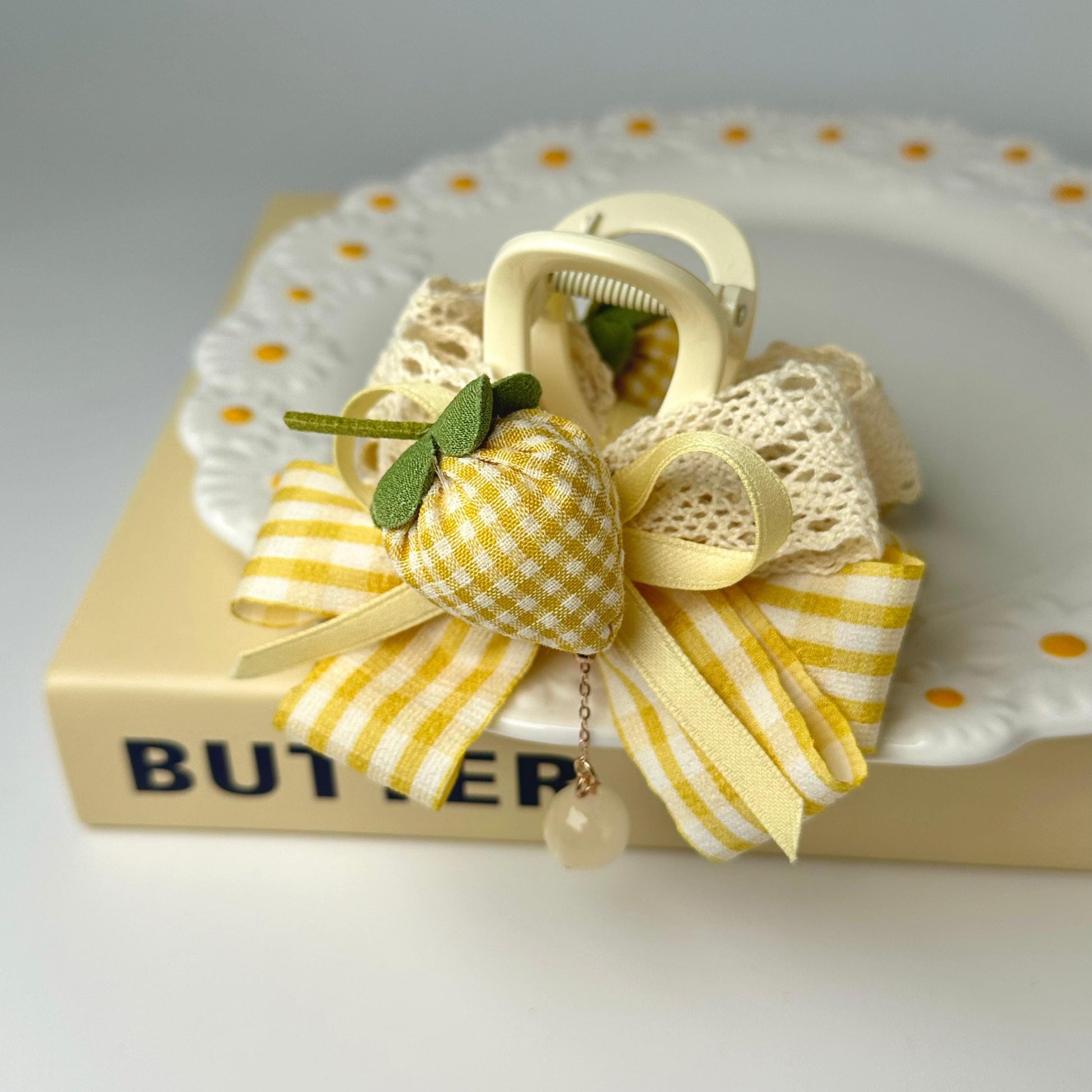 A decorative bow made of fabric and ribbons, featuring a yellow and white checkered pattern, is placed on top of a white plate with a scalloped edge.