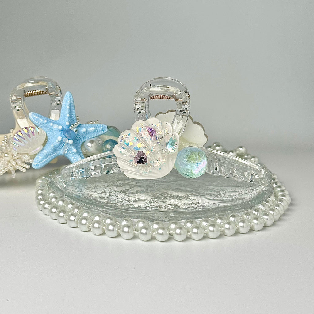 A clear glass hair clip adorned with seashells, pearls, and a starfish, set against a white background.