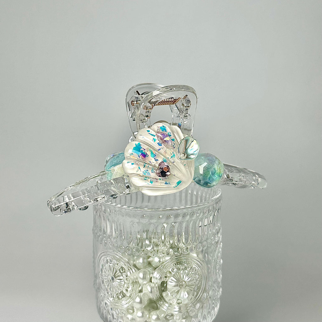 A clear glass container filled with small, round, multicolored pearls, with a clear plastic object featuring a shell-shaped design and colorful accents placed on top of the container.