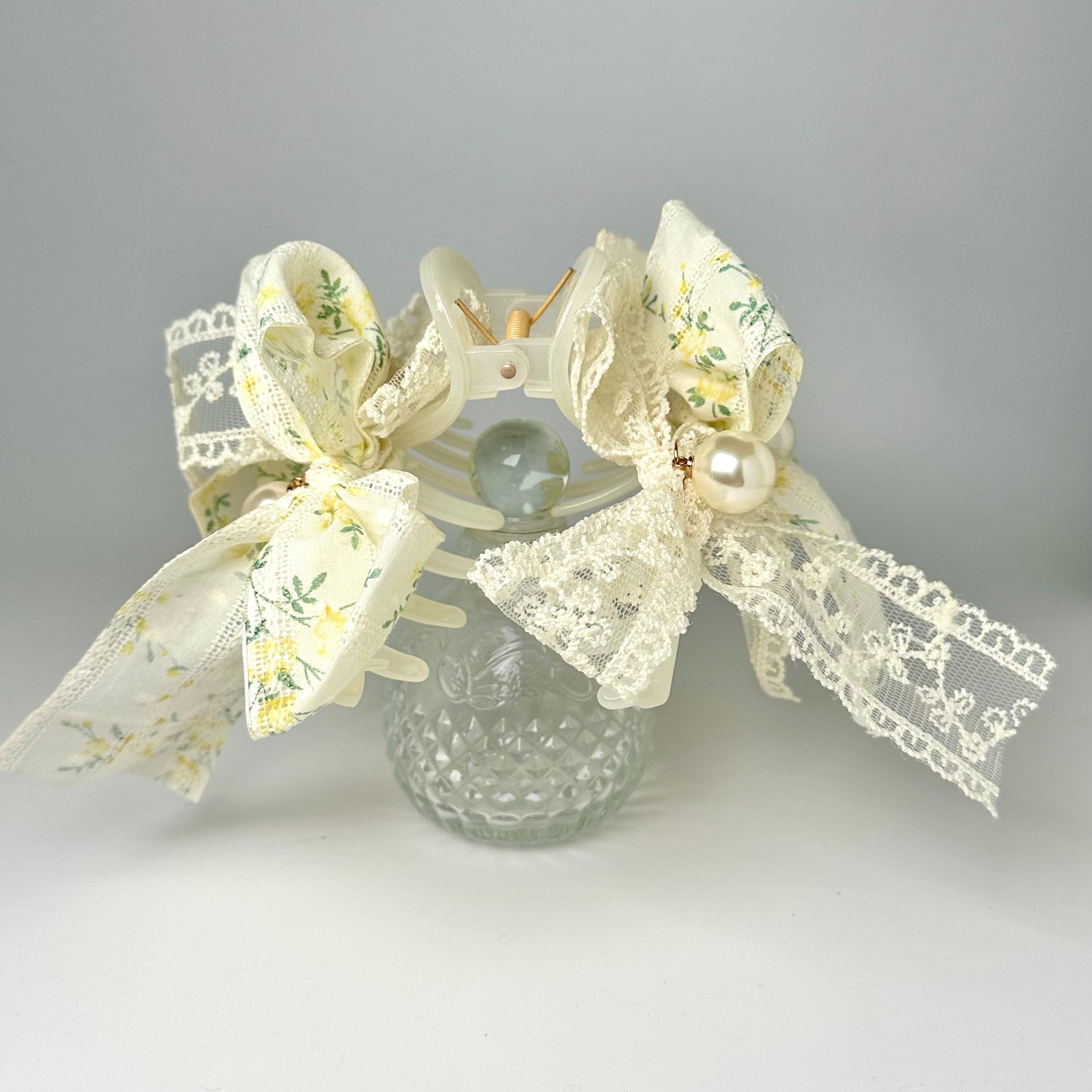 A glass vase with a floral pattern and lace trim, adorned with delicate bows and pearls, sits on a white surface.
