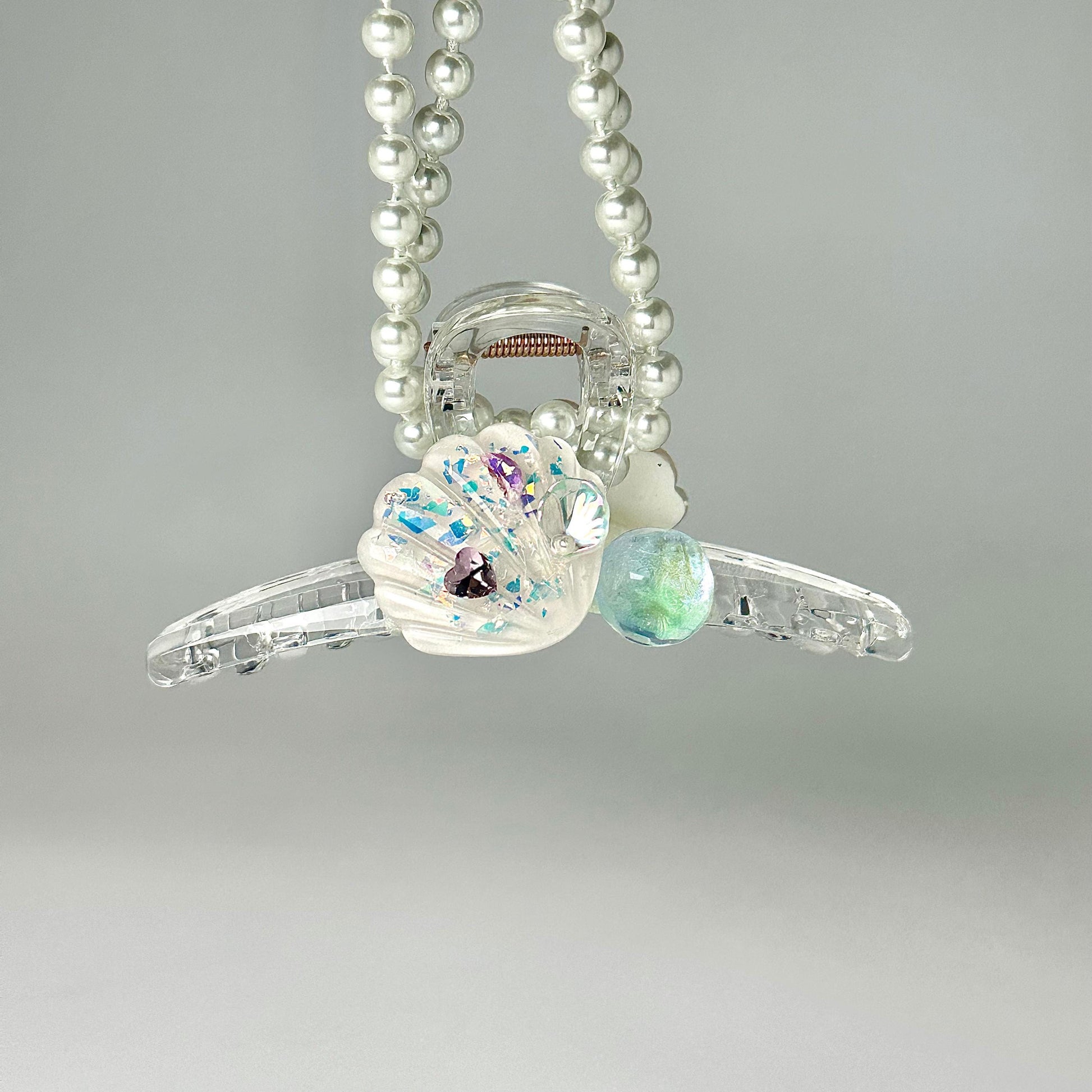 A necklace with clear beads and a shell-shaped pendant, adorned with colorful gemstones, is suspended against a light gray background.