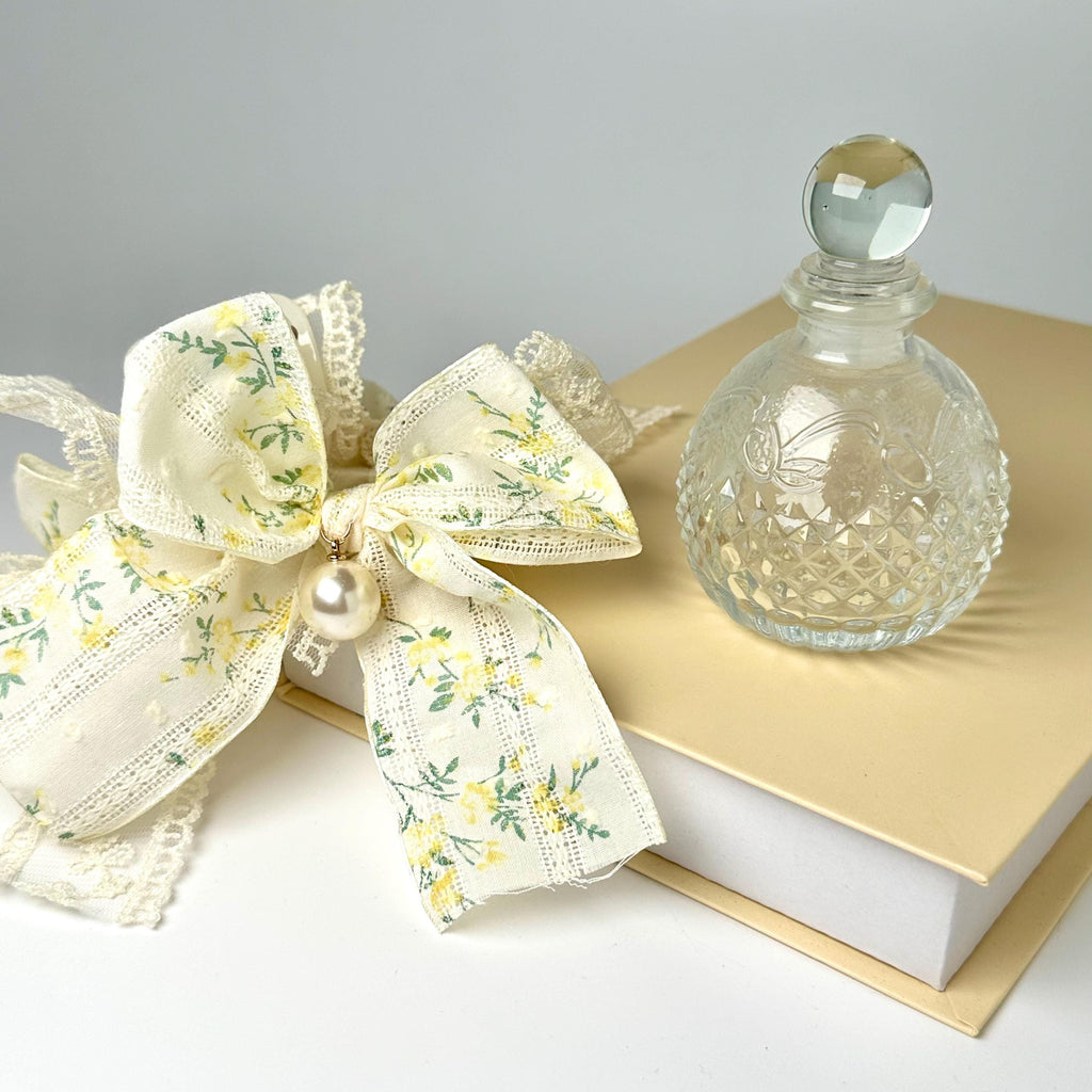 a clear glass perfume bottle with a white lace bow and a white book with a gold ribbon on top, all placed on a white surface.