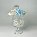 A clear glass vase filled with small white pearls and a blue starfish is placed on a white surface.