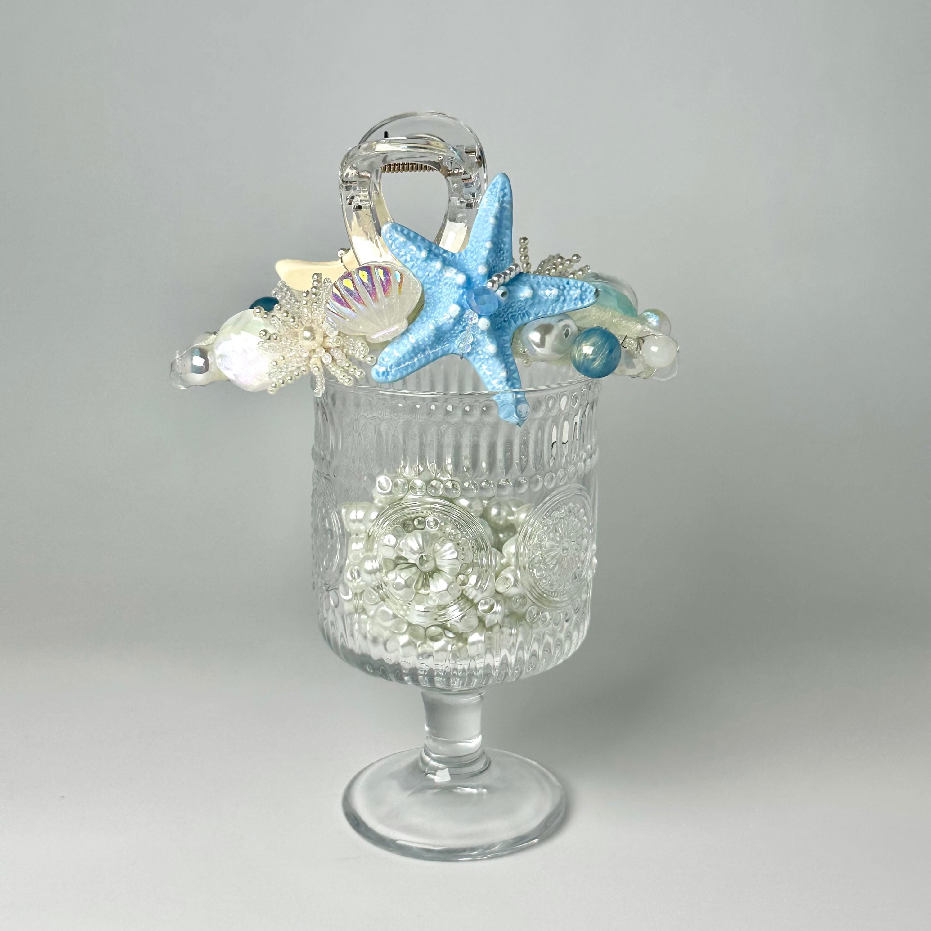 A clear glass vase filled with small white pearls and a blue starfish is placed on a white surface.