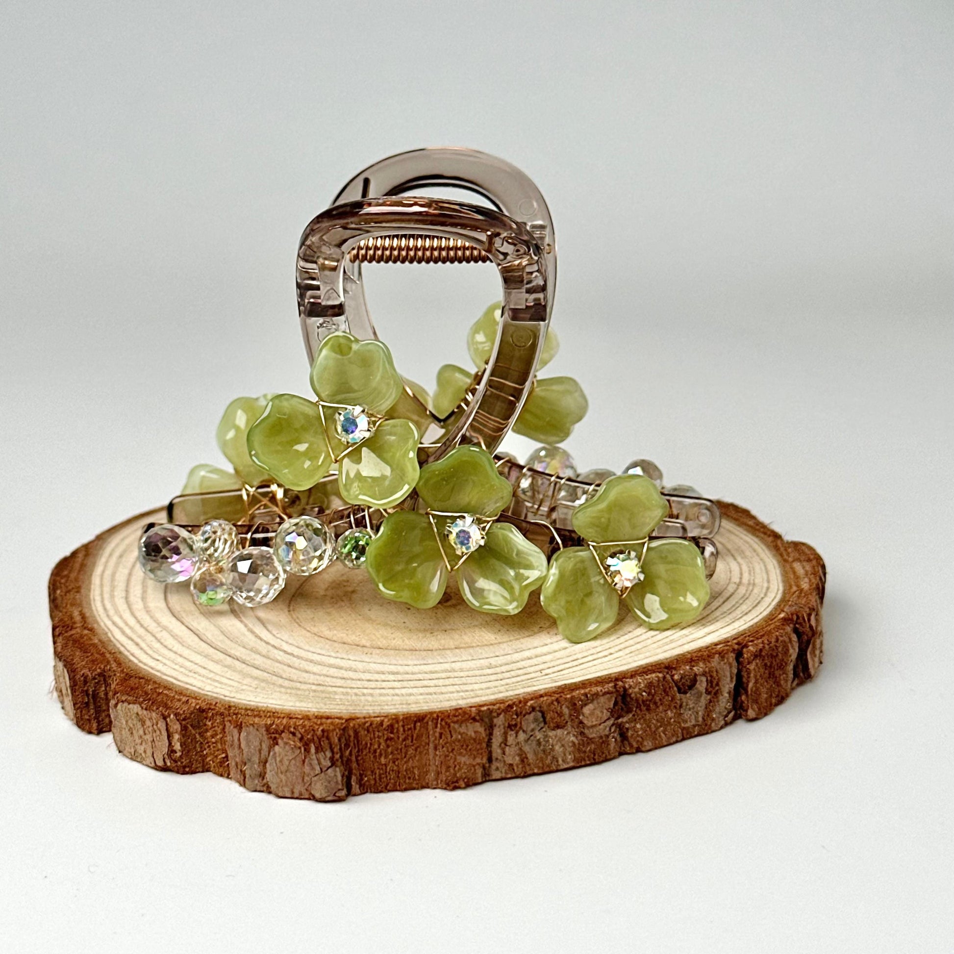 A silver hair clip adorned with green glass flowers and rhinestones is displayed on a wooden slice.