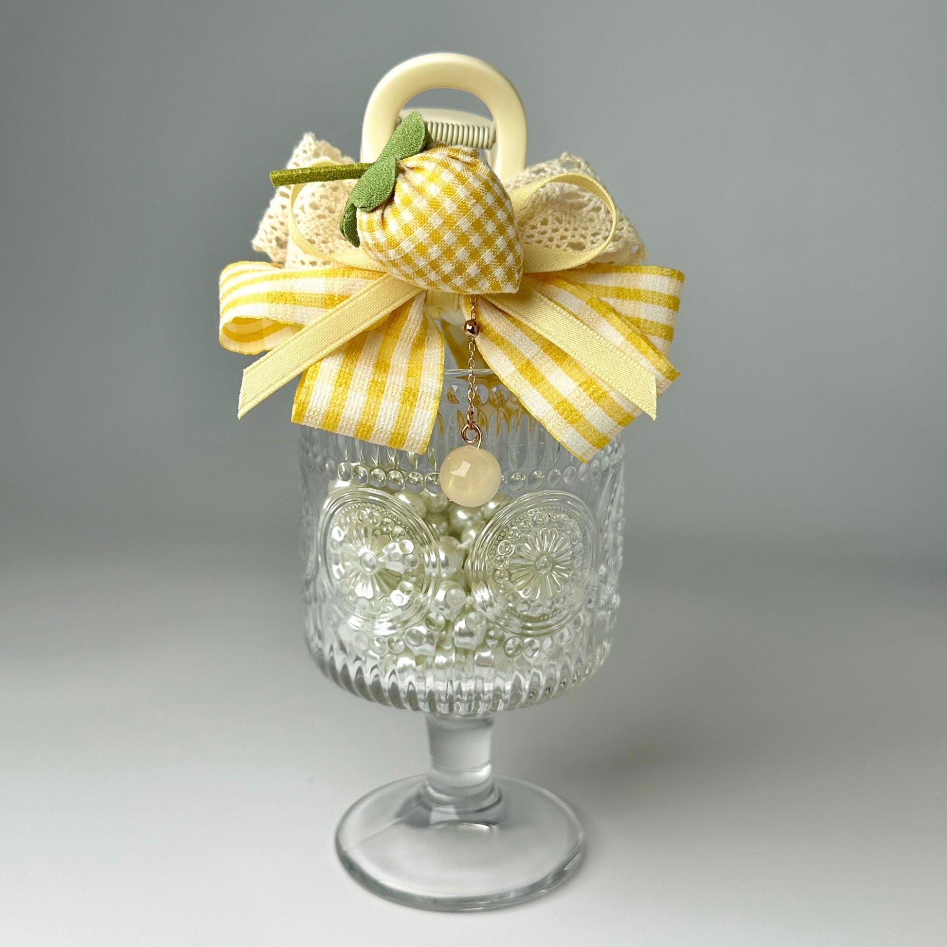 A glass vase with a yellow and white checkered bow, a small charm, and a white flower, placed on a white surface.