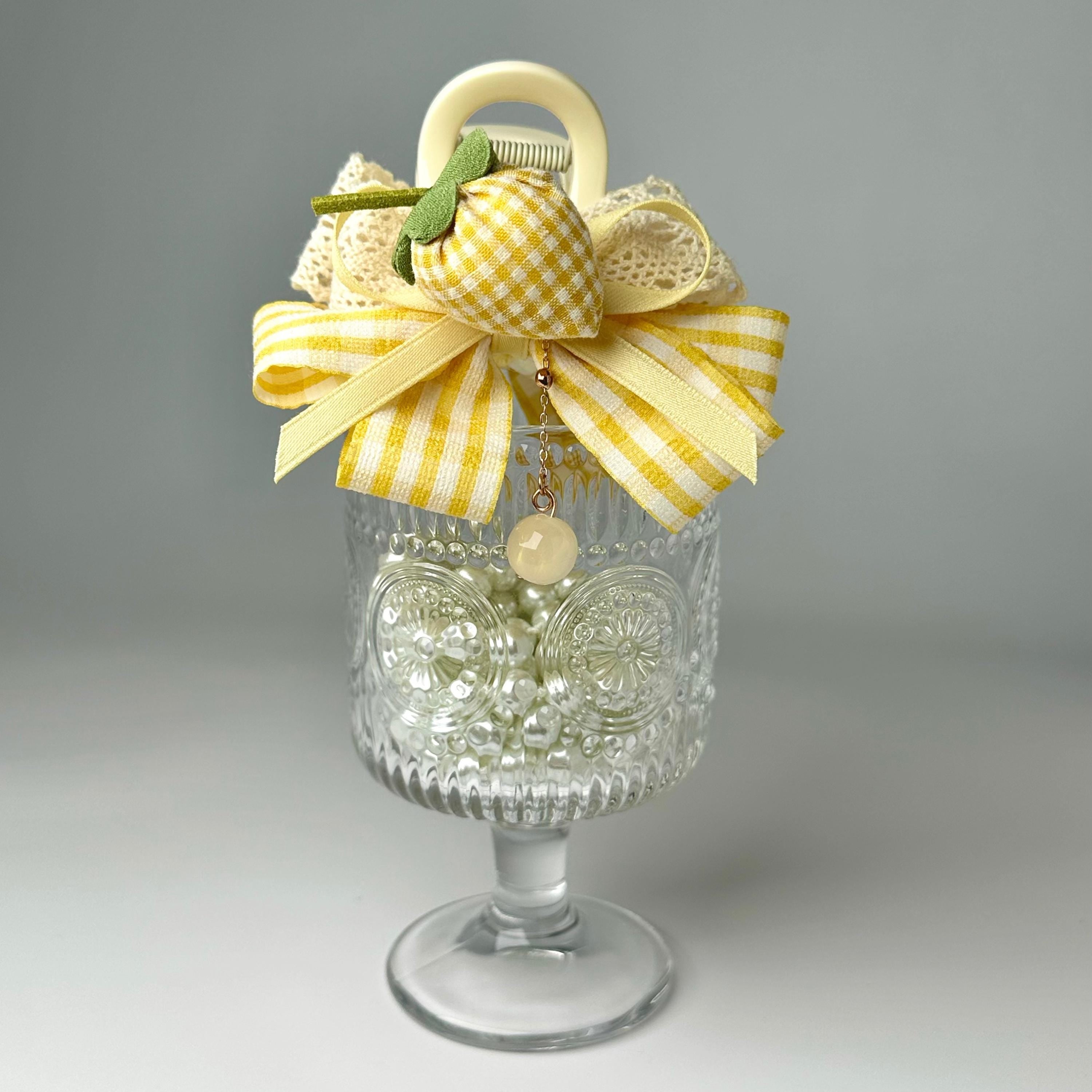 A glass vase with a yellow and white checkered bow, a small charm, and a white flower, placed on a white surface.